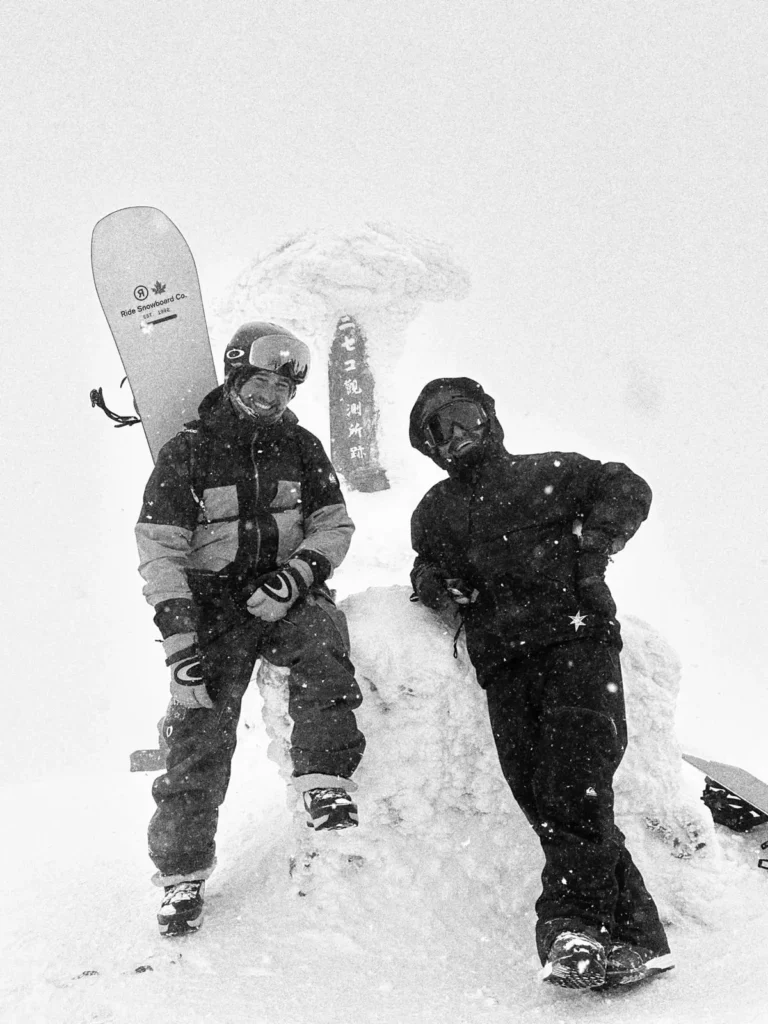 Two snowboarders, one carrying a powder board, beside the frozen summit shrine of Mount Niseko Annupuri in a snowstorm